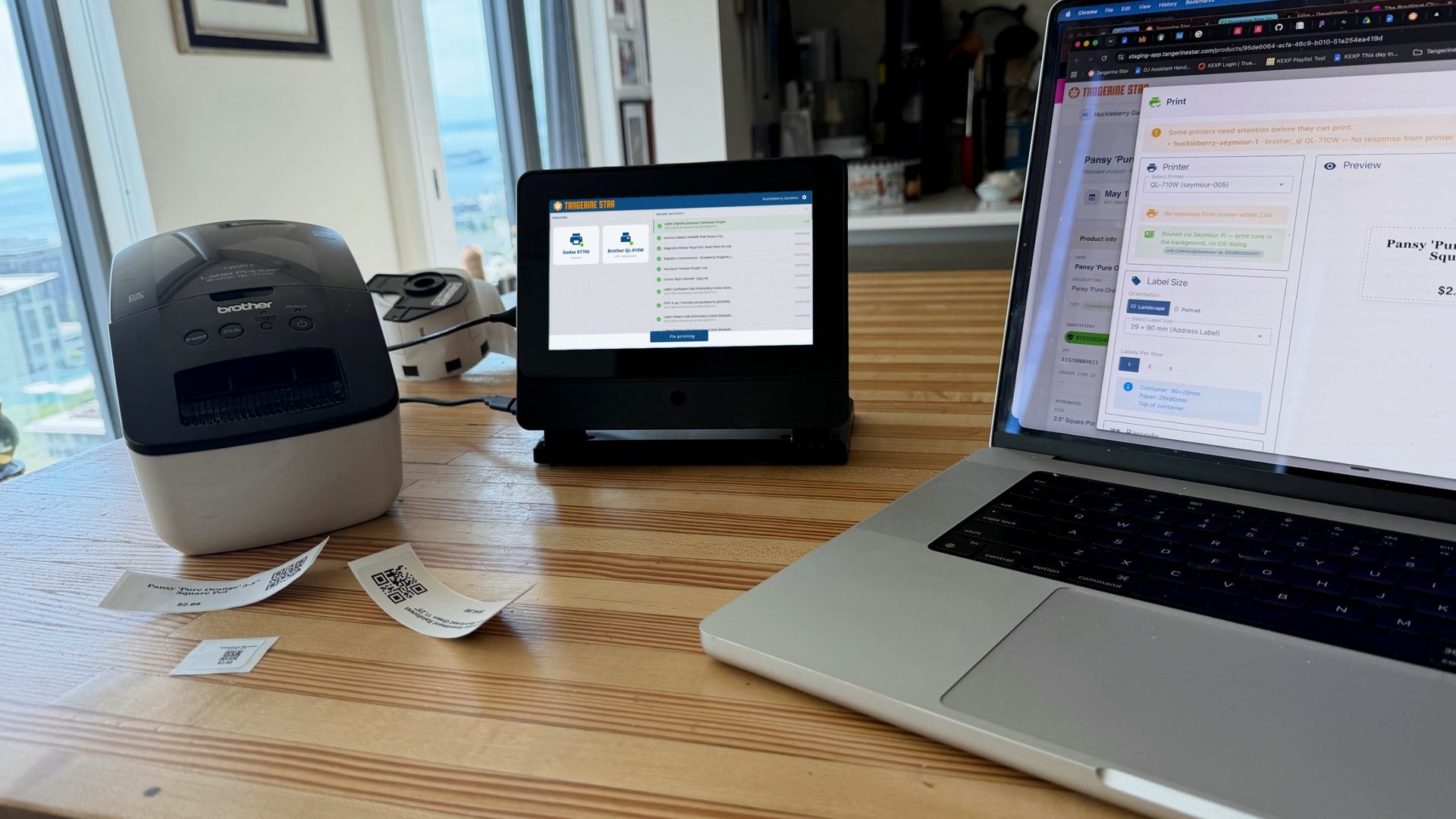 Seymour on a shop counter showing the live label-print dashboard, beside a Brother label printer and freshly printed QR labels